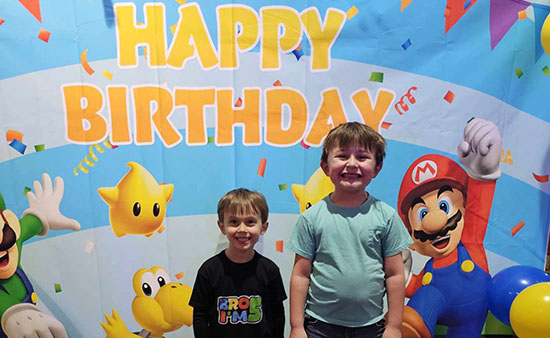 Two children stand in front of a colorful birthday backdrop featuring Mario characters and "Happy Birthday" text.