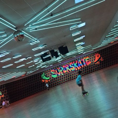 A vibrant roller rink with neon lights and a disco ball, featuring a colorful "SUPERSKATE" mural and skaters gliding on the floor.