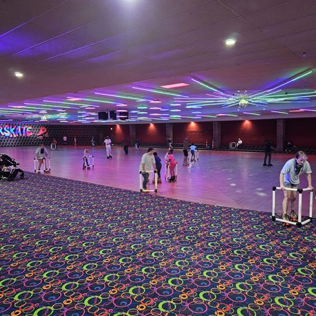 Brightly lit roller rink with colorful lights and a playful atmosphere, featuring skaters of all ages, including children with skating aids.