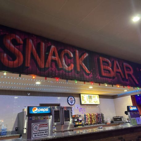 Brightly lit snack bar with a colorful sign, beverage dispensers, and assorted snacks displayed on a counter.