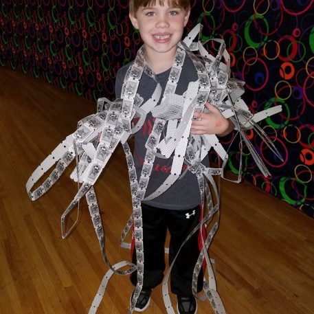 A child stands on a wooden floor, playfully wrapped in long strips of paper tickets, surrounded by a colorful patterned background.