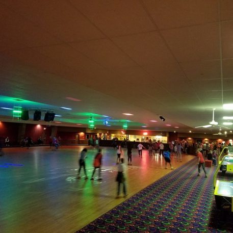 A lively roller skating rink filled with skaters under colorful lights, featuring arcade games in the foreground and a snack bar in the background.