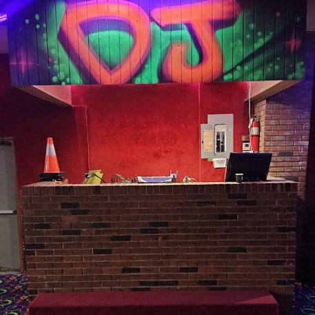A brightly lit DJ booth features graffiti-style "DJ" lettering above a brick counter, with colorful patterned flooring and a fire extinguisher nearby.
