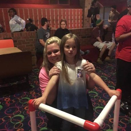 Two children pose playfully behind a railing in a vibrant bowling alley filled with colorful carpet, while others socialize in the background.