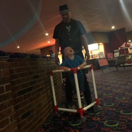 A person in a black outfit pushes a child in a homemade cart made of white and red plastic pipes, inside a brightly colored venue.