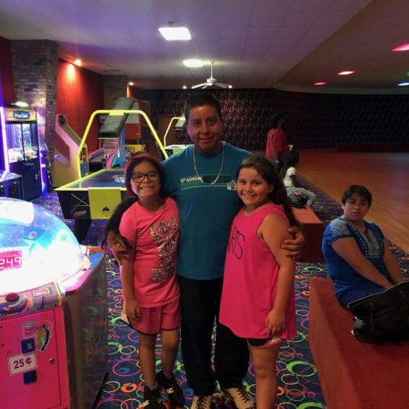 A man poses with two young girls in roller skates at a brightly colored skating rink, featuring arcade games in the background.