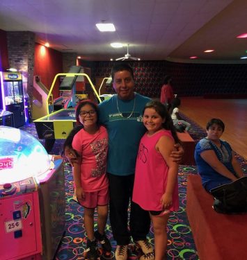Two girls and a man stand together in front of a brightly lit bowling alley entrance.