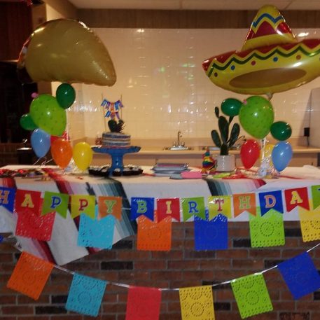 Colorful birthday decorations featuring sombrero balloons, a cake, and a "Happy Birthday" banner on a festive tablecloth.
