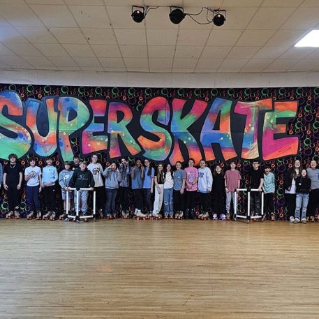 A group of kids in skate shoes poses in front of a colorful "SUPERSKATE" mural in a roller skating rink.