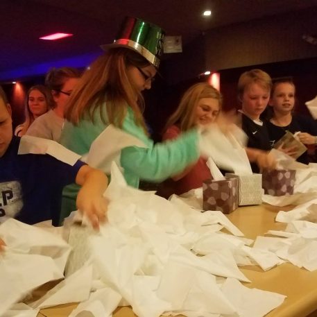 A group of children excitedly unwrap gifts, surrounded by scattered tissue paper and gift boxes on a table in a festive setting.
