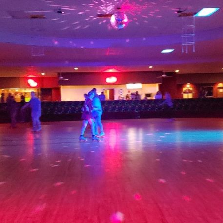 A vibrant roller rink with colorful lights, featuring couples skating and a disco ball reflecting patterns on the wooden floor.