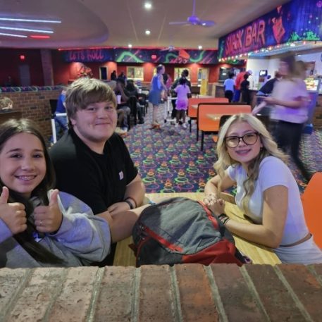 A group of friends poses at a social venue, with vibrant decor and a colorful carpet, while others engage in various activities around them.