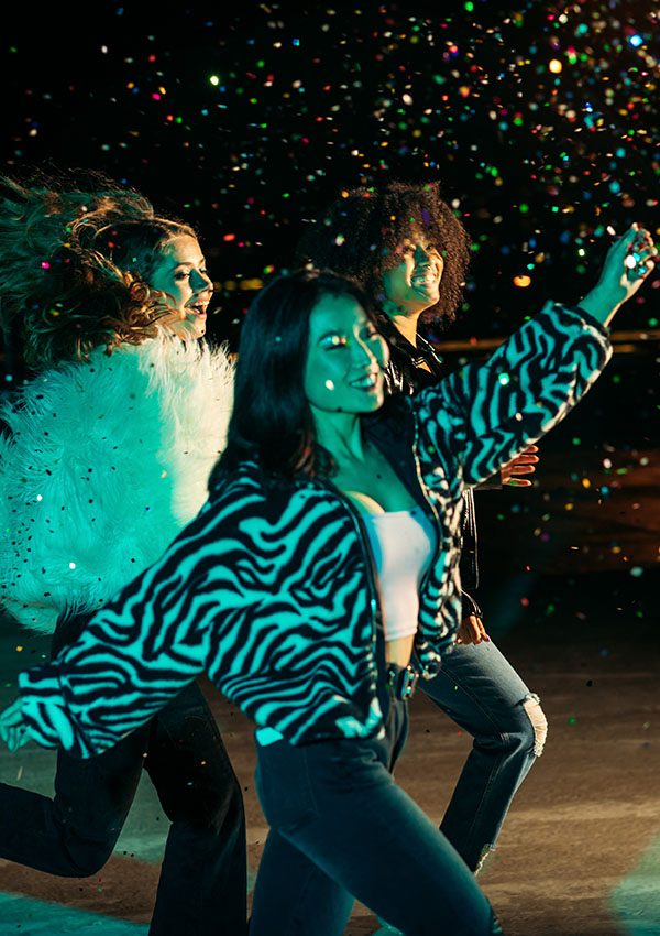 Three individuals dance joyfully under colorful, sparkling confetti, wearing stylish outfits in a vibrant nightlife setting.
