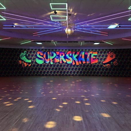 Interior of a roller skating rink named "Superskate," featuring colorful neon lights, a disco ball, and a vibrant graffiti-style mural.