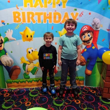 Two children on roller skates pose in front of a colorful "Happy Birthday" backdrop featuring cartoon characters and balloons.