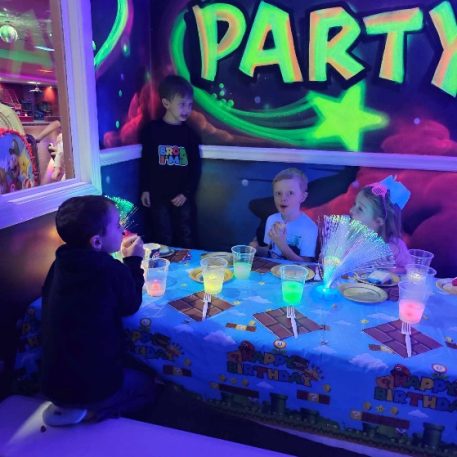 A colorful party scene with children seated around a decorated table, enjoying drinks and snacks under vibrant, glowing decorations.