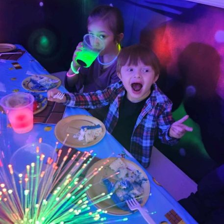 Two children at a colorful party table decorated with blue cake and glowing drinks, surrounded by vibrant light effects.