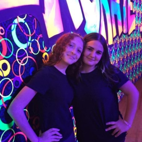 Two friends pose together in front of a vibrant, neon-lit wall featuring colorful patterns and the word "SKATE."