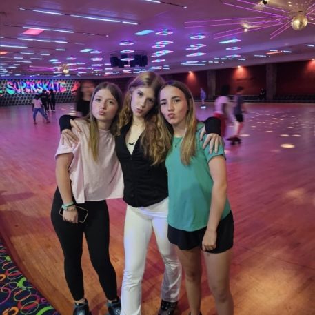 Three friends pose together in a roller skating rink, wearing colorful outfits and skates, with vibrant lights illuminating the space.