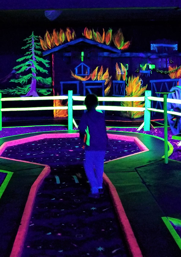 A child walks along a glowing mini-golf course surrounded by vibrant, neon artwork of a burning house and trees.