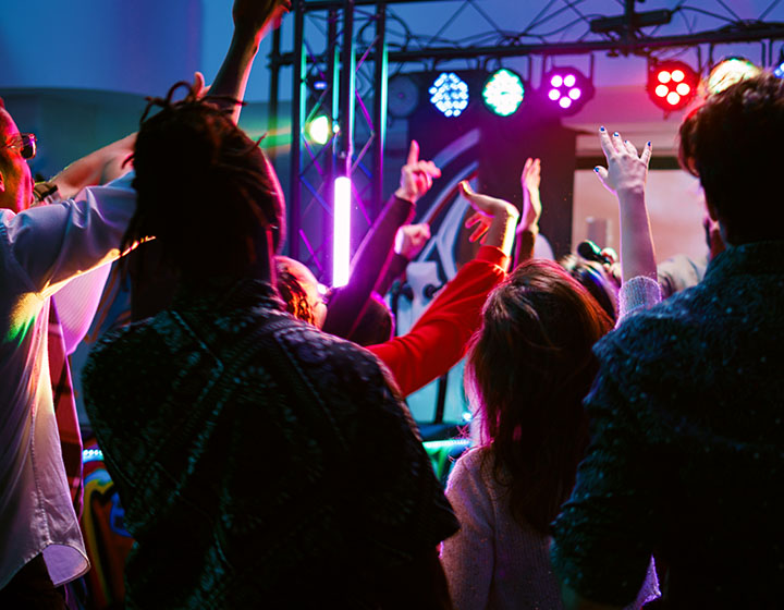 A lively crowd dances with raised hands under colorful lights at a vibrant event.