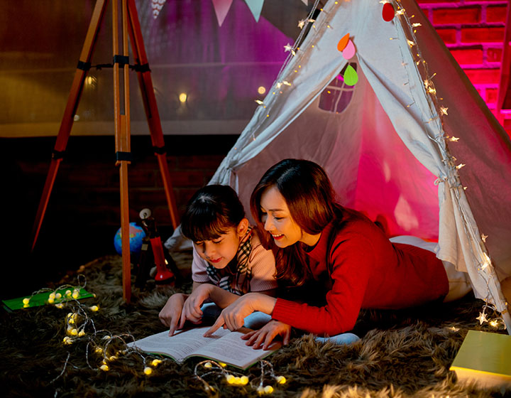A cozy indoor scene with two people reading a book inside a tent, surrounded by fairy lights and a warm, inviting atmosphere.